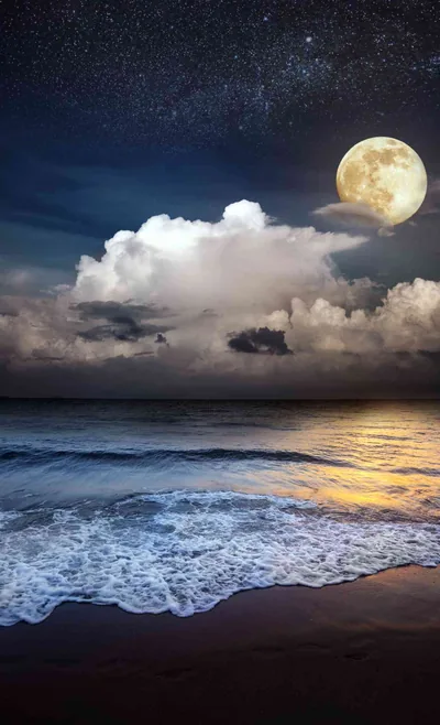A bright full moon sits above a bank of cloud, its light running in a broken column down to the breaking surf.