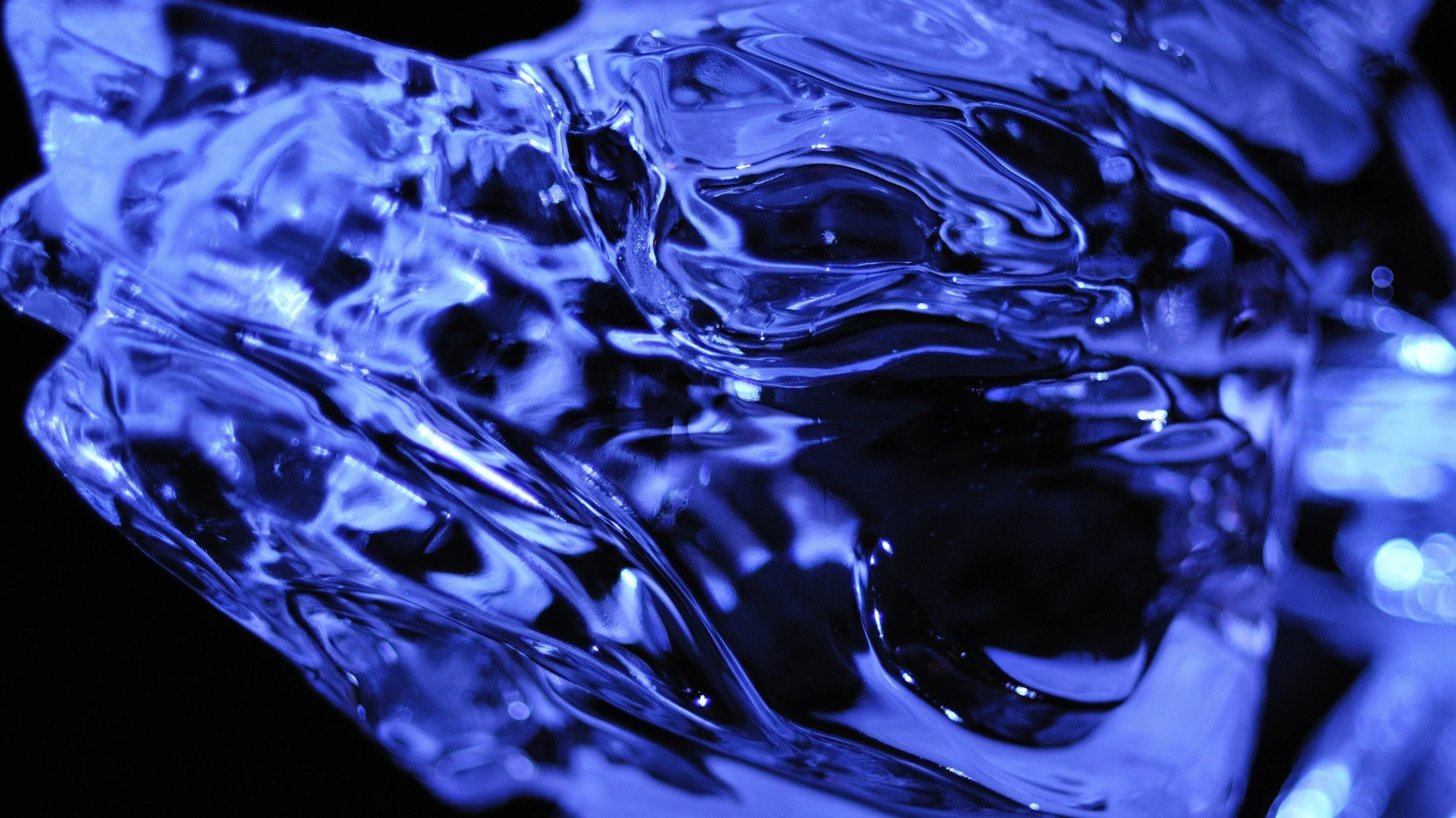A macro view of blue liquid catching hard light, its ridges and bubbles glassy and sharp while the background drops into shadow.