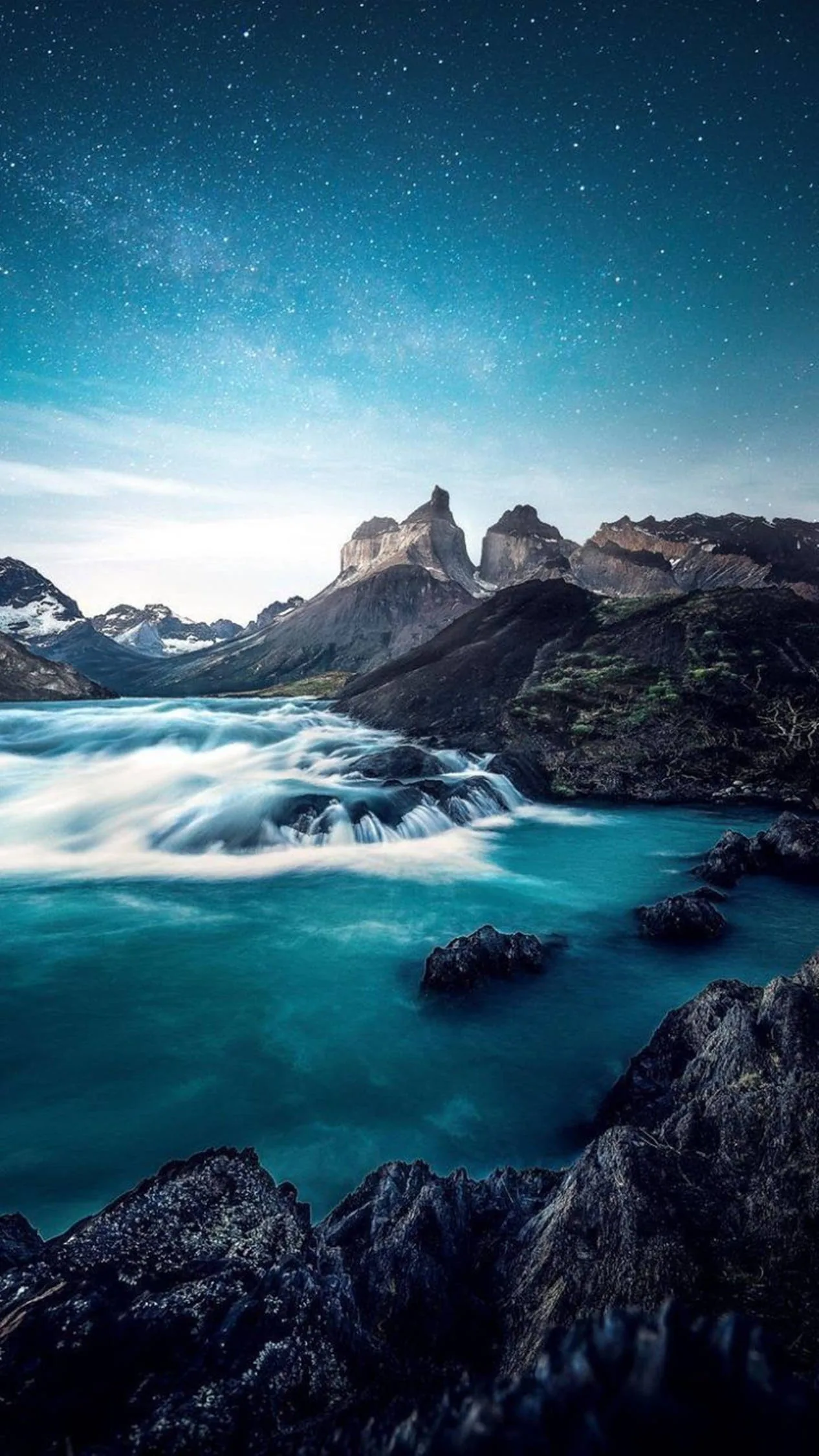 A milky turquoise river runs between dark boulders toward sharp granite peaks, with a star-scattered blue sky filling the upper half.