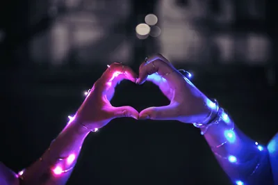Two hands wrapped in string lights meet to form a heart, pink on the left and blue on the right with bokeh scattered behind.