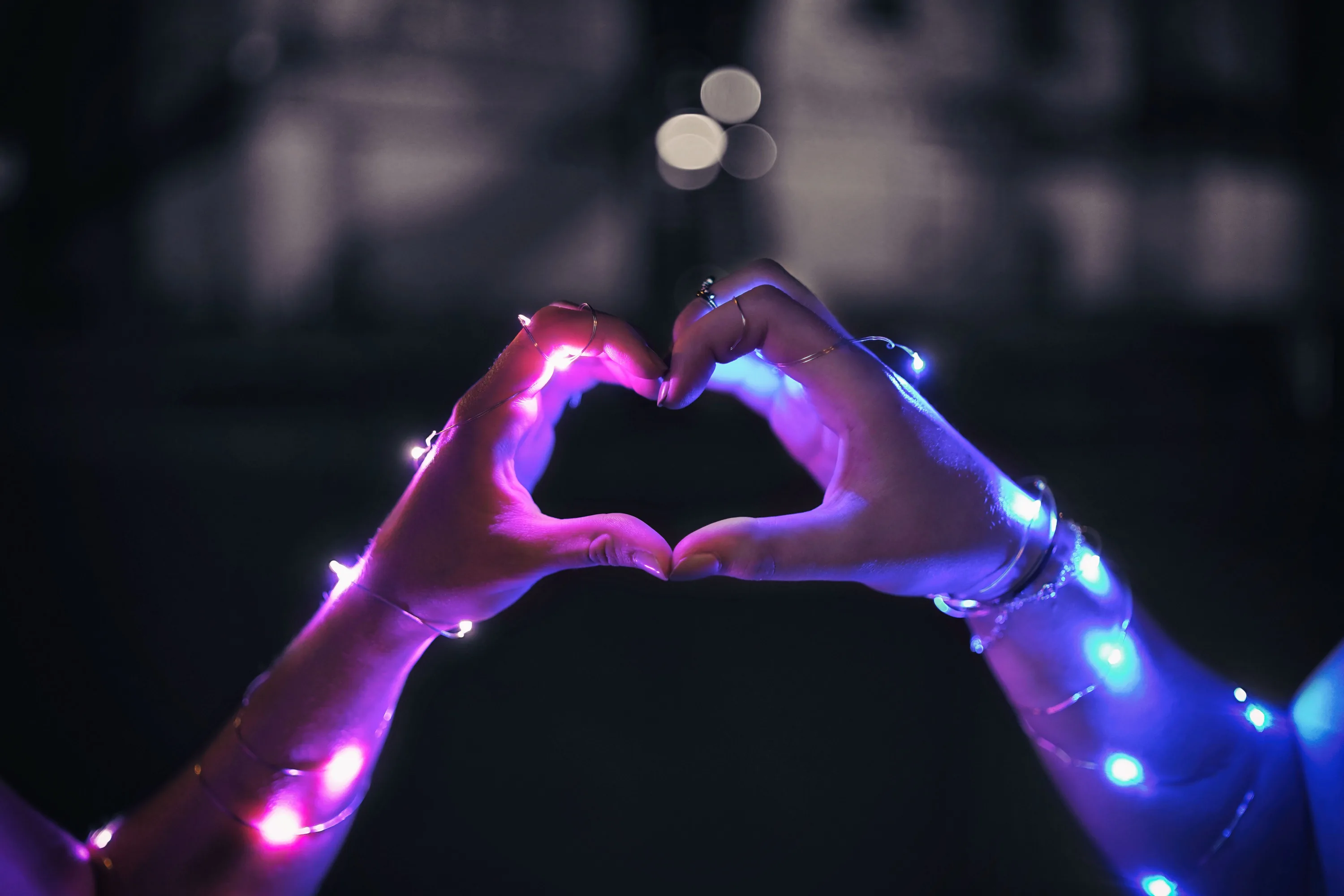 Two hands wrapped in string lights meet to form a heart, pink on the left and blue on the right with bokeh scattered behind.