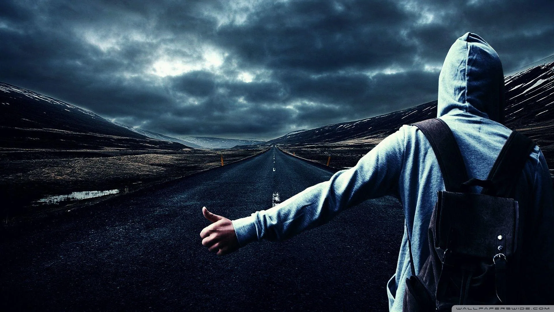 Seen from behind, a boy in a grey hoodie with a backpack sticks his thumb out on a rain-slicked road running into stormy hills.