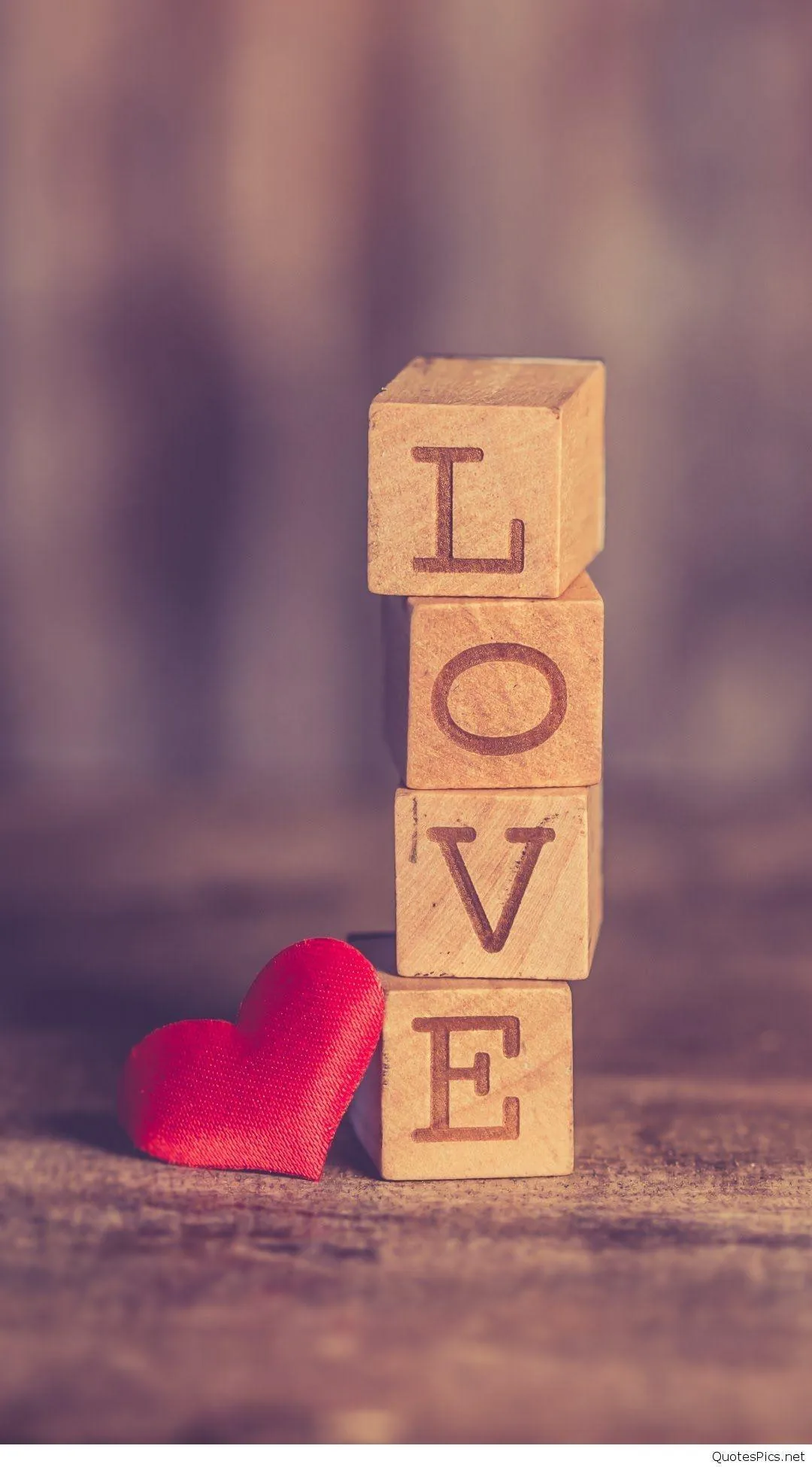 Four lettered blocks stack into LOVE on a worn wood table, a soft red fabric heart leaning at the base in warm light.