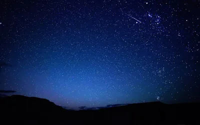 Two thin meteor trails cross near a bright star cluster at the top right, above hills that fade to full black along the bottom edge.