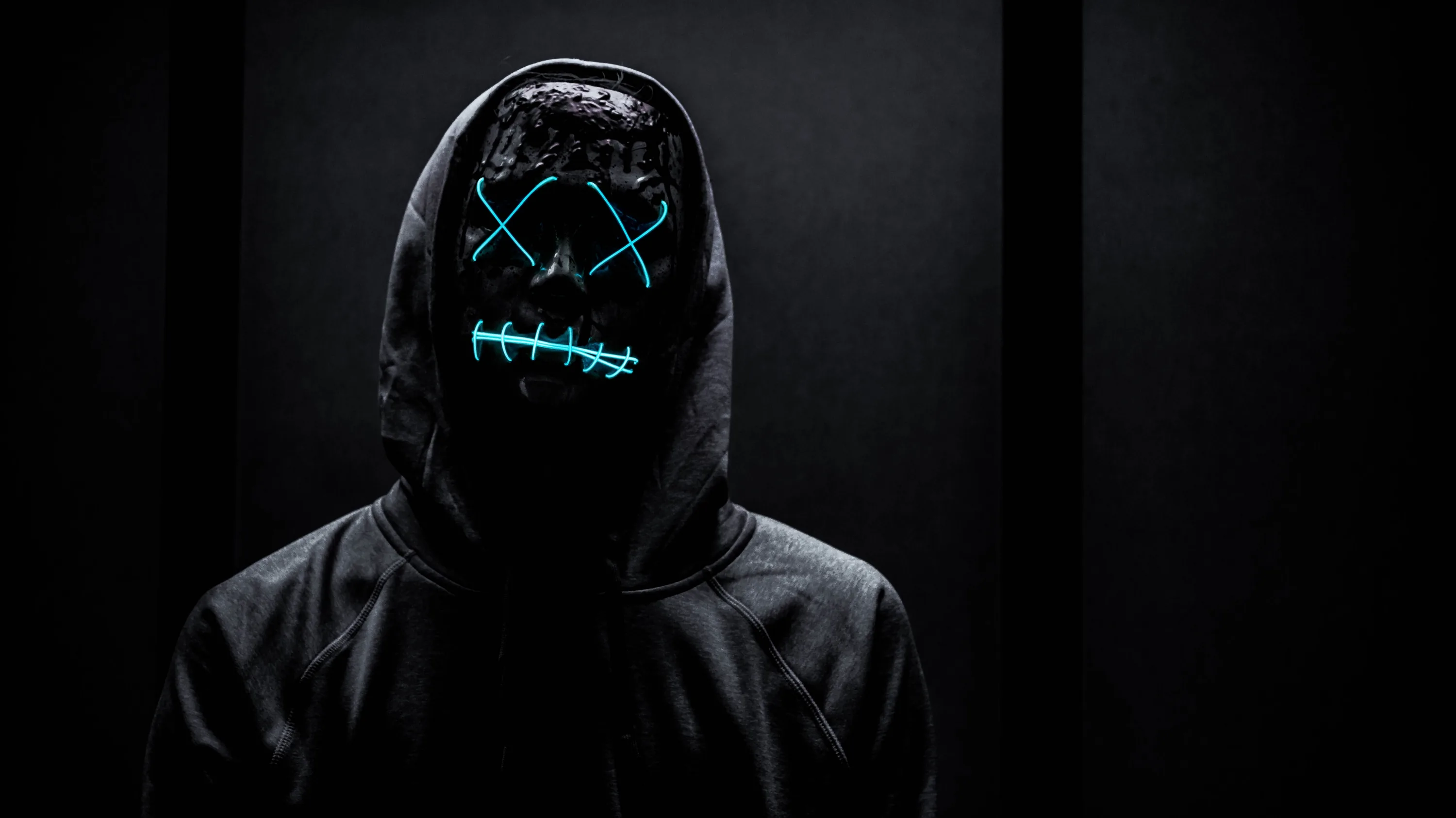 A figure in a black hoodie faces the camera, cyan stitched lines across the mask the only light in an otherwise grey-black frame.