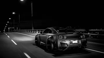 Rear view of a wide-arch GT-R pulling away down an elevated road, streetlights receding on the left and the whole scene rendered in grainy black and white.