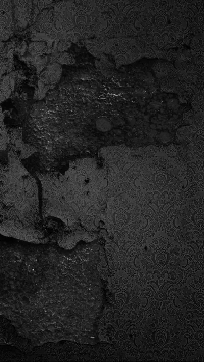 Old black paint flakes away from a concrete wall in rough patches, side light picking out the grit where the surface has broken open.