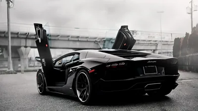Both doors stand vertical on an Aventador parked beneath a bridge, the open cabin and forged wheels lit by flat overcast light on damp tarmac.