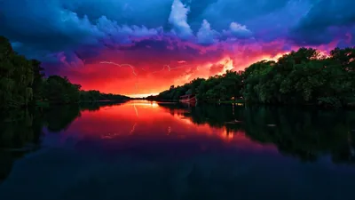 Forked lightning drops through purple storm clouds dead centre, the river below returning the whole sky as a mirror; both dark tree lines frame the glow.