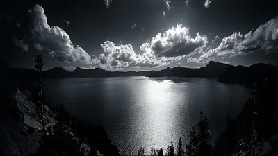 Cloud banks pile above a wide lake in monochrome, moonlight breaking into a bright column down the water.