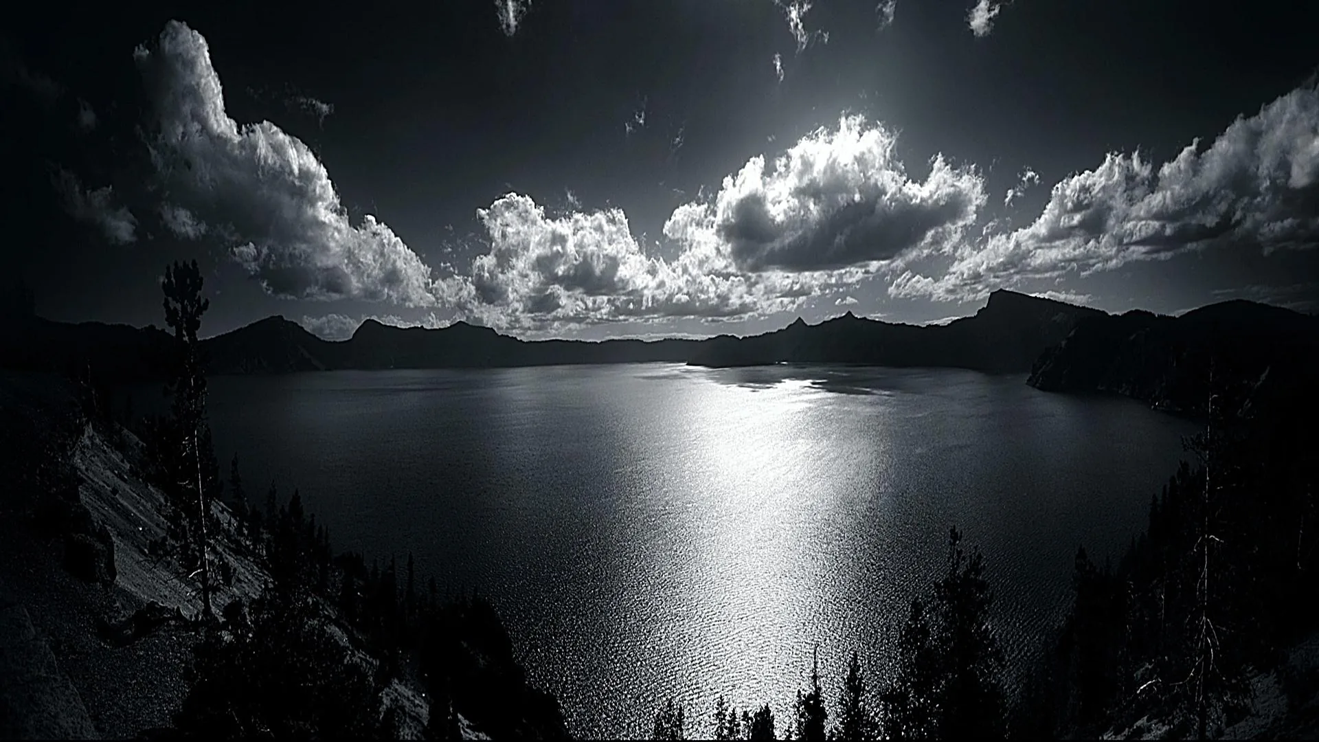 Cloud banks pile above a wide lake in monochrome, moonlight breaking into a bright column down the water.