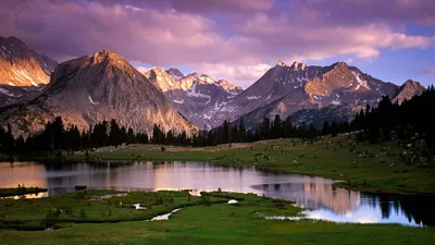 A stream winds through a grassy basin below granite peaks under lavender clouds, low sun catching the left-hand slopes.