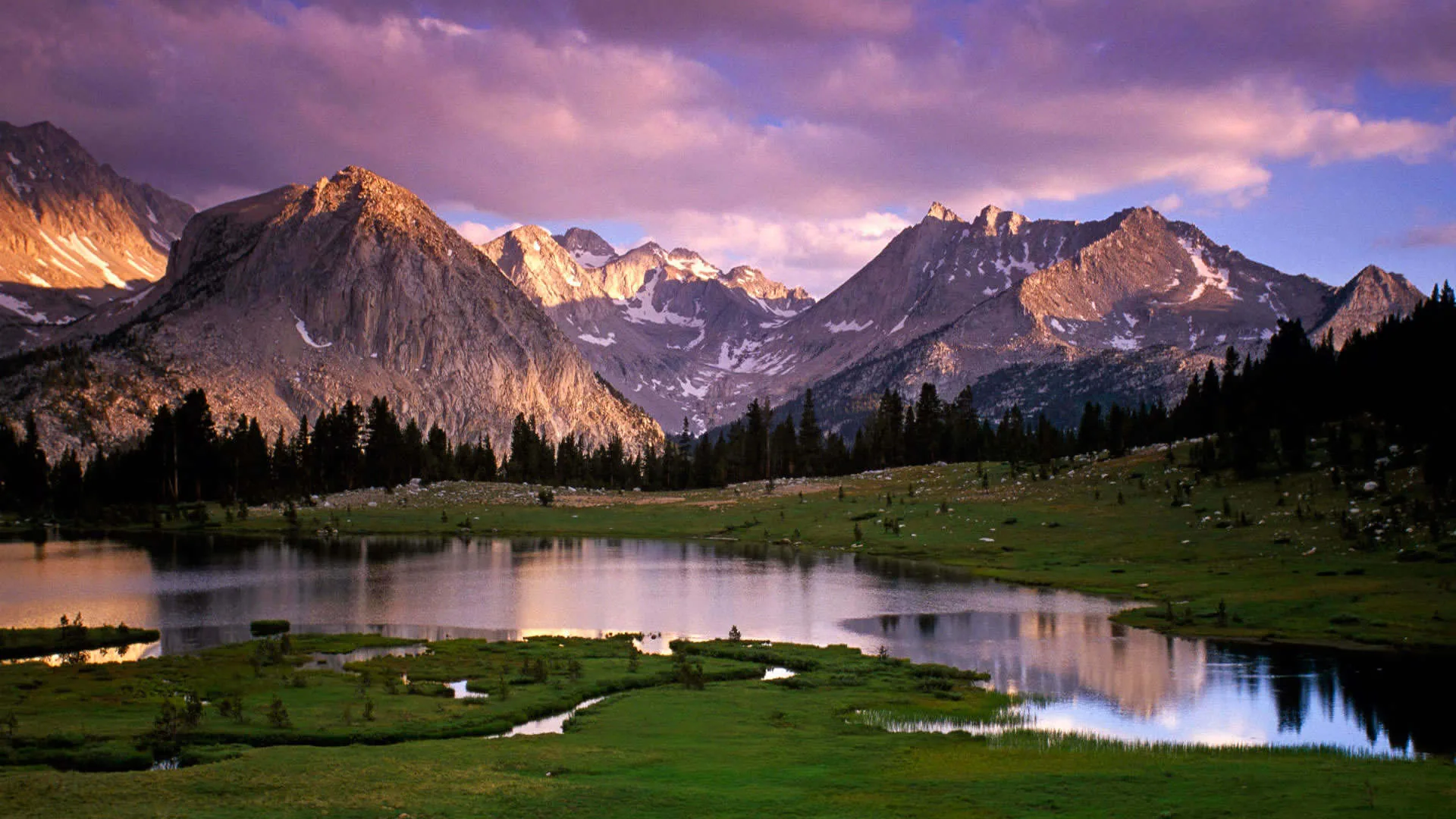 A stream winds through a grassy basin below granite peaks under lavender clouds, low sun catching the left-hand slopes.