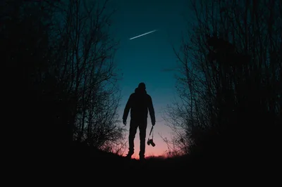 A single meteor streak crosses the teal sky between two dark tree lines, the man below holding a camera as orange dusk burns at the horizon.
