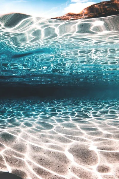 Shot half beneath the surface, sunlight weaves bright ripple patterns across the sand while a rock ledge shows above the waterline.