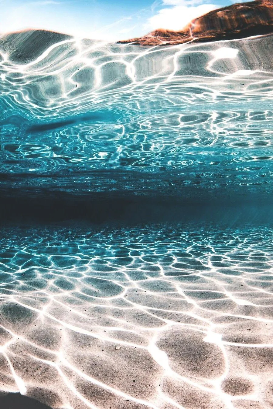 Shot half beneath the surface, sunlight weaves bright ripple patterns across the sand while a rock ledge shows above the waterline.
