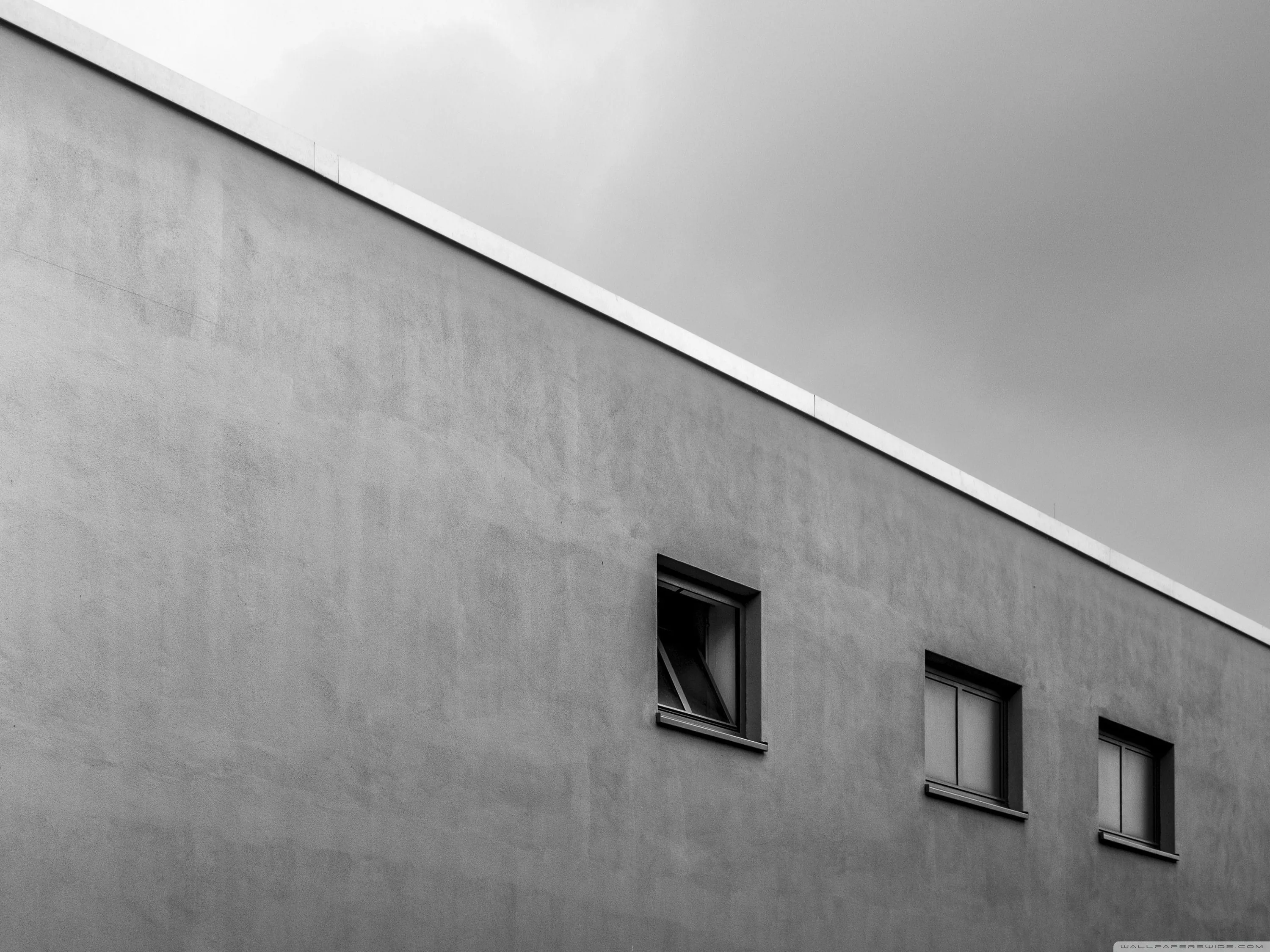 The roofline of a plain rendered building cuts a diagonal across an overcast sky, three small square windows punched into the wall below.