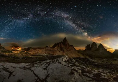 The Milky Way arches over jagged Dolomite peaks in a stitched panorama, a lit cabin glowing orange at the left edge of the valley.