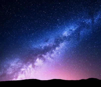 The galactic band crosses a starry sky above black hills, pink light pooling along the horizon and fading to violet higher up.