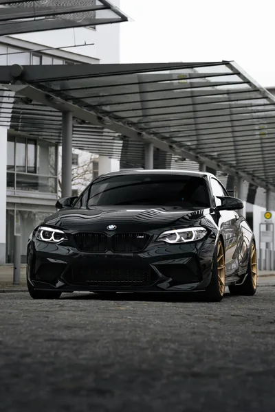The M2 faces the camera under a ribbed steel canopy, its dark kidney grille and angel-eye headlamps set against pale office blocks in the distance.