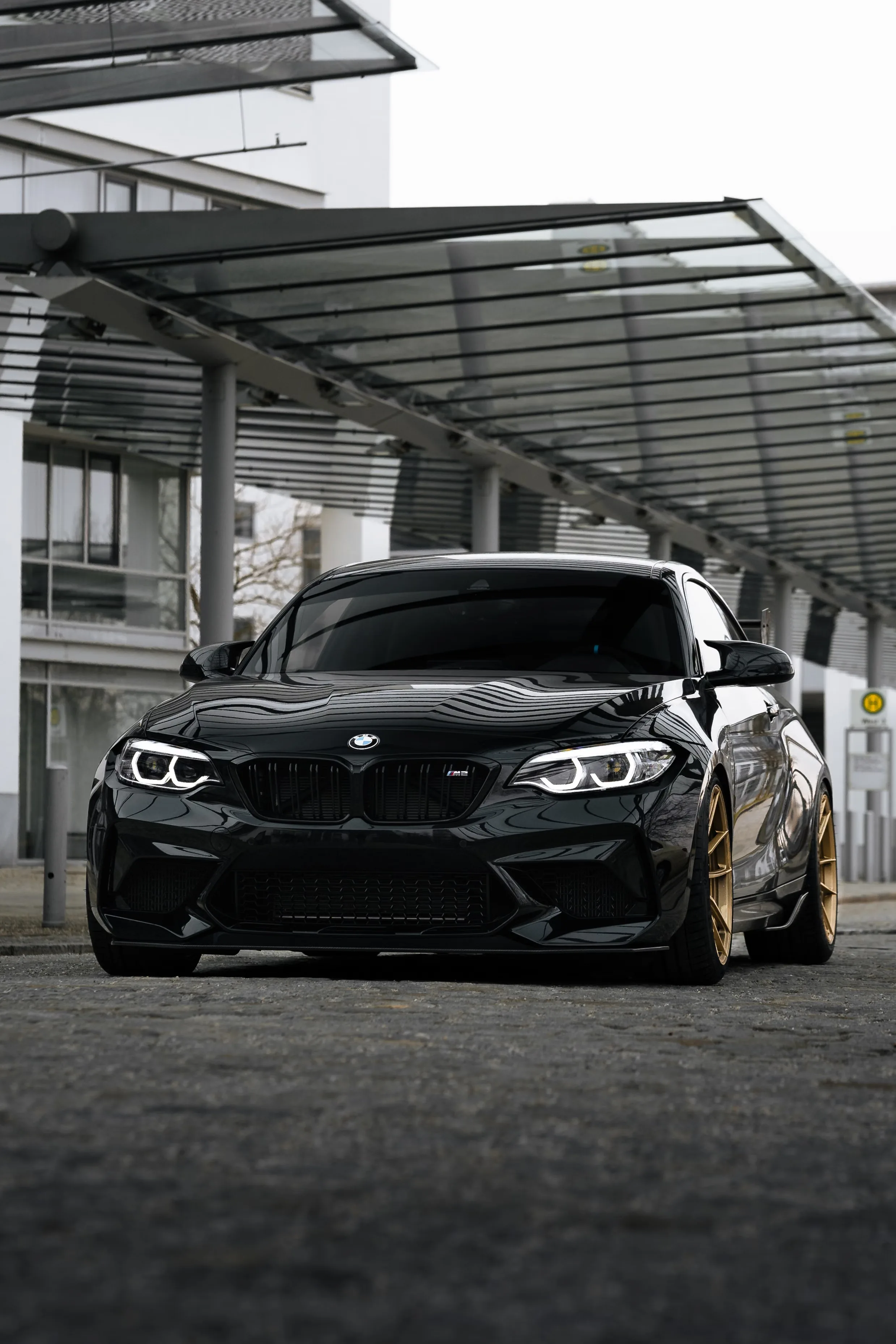The M2 faces the camera under a ribbed steel canopy, its dark kidney grille and angel-eye headlamps set against pale office blocks in the distance.