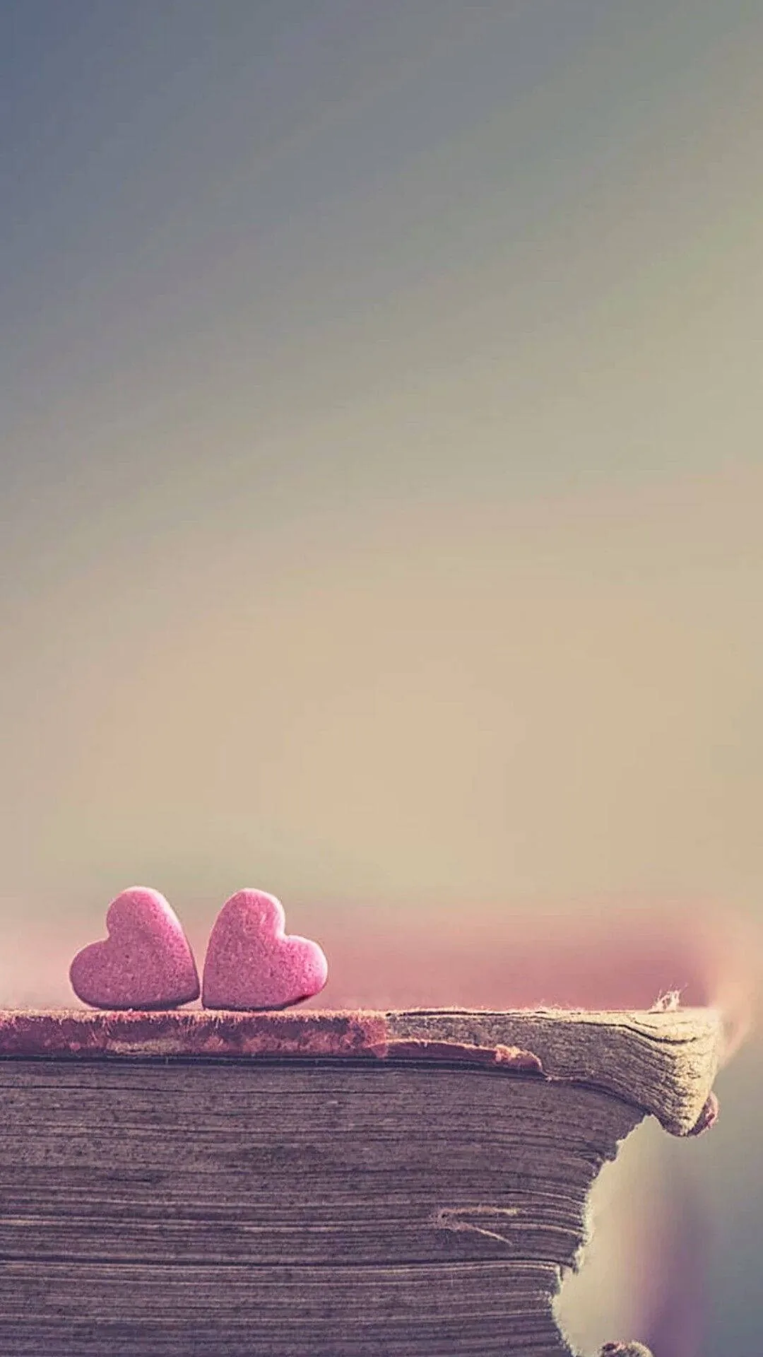 Two small pink candy hearts sit on the frayed edge of an old book, the background washed out to pale teal and cream.