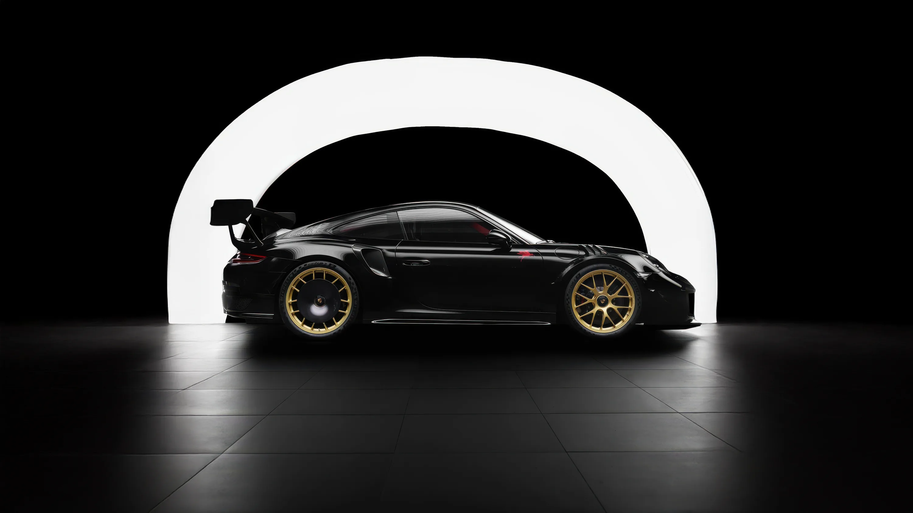 A black Porsche race car in dead side profile, gold centre-lock wheels and a tall rear wing outlined by a glowing white arch on an otherwise black wall.