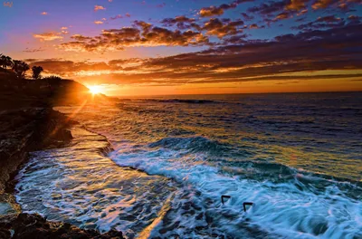The sun bursts from behind a low headland and lights streaks of foam across churning water, dark rock ledges holding down the left corner.