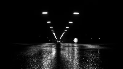 Black and white: overhead lights recede down a wet road, each one doubled in the reflection on the tarmac.