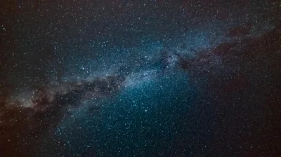 The Milky Way crosses the frame diagonally in teal and dust-brown, dense with stars yet dark enough at the corners for a desktop layout.