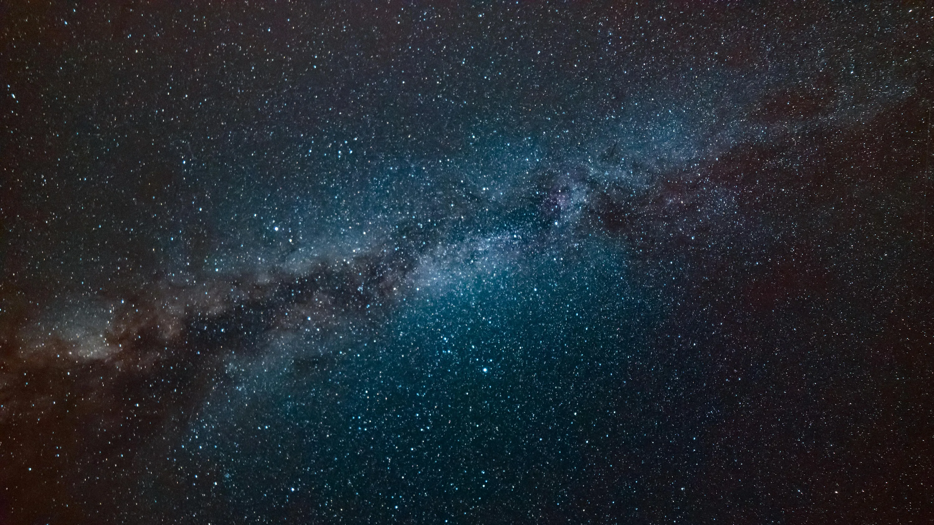 The Milky Way crosses the frame diagonally in teal and dust-brown, dense with stars yet dark enough at the corners for a desktop layout.