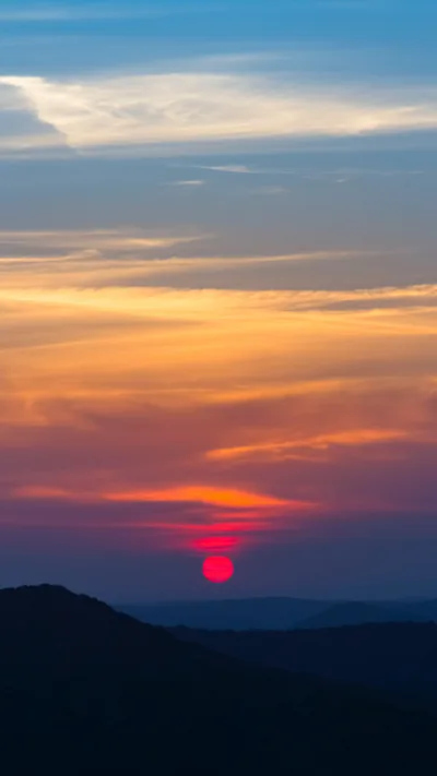 A small red sun drops behind layered dark hills, an orange band across the middle fading to blue-grey at the top where a clock stays readable.