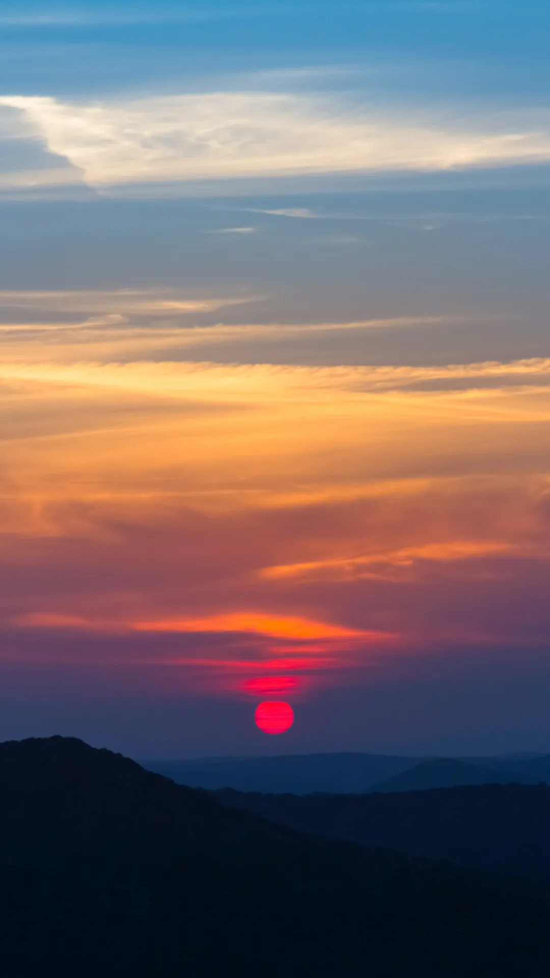 A small red sun drops behind layered dark hills, an orange band across the middle fading to blue-grey at the top where a clock stays readable.
