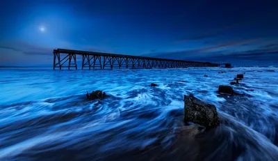 A long exposure at blue hour, tide streaming over dark rocks toward a ruined loading pier, with the moon small in the upper left sky.