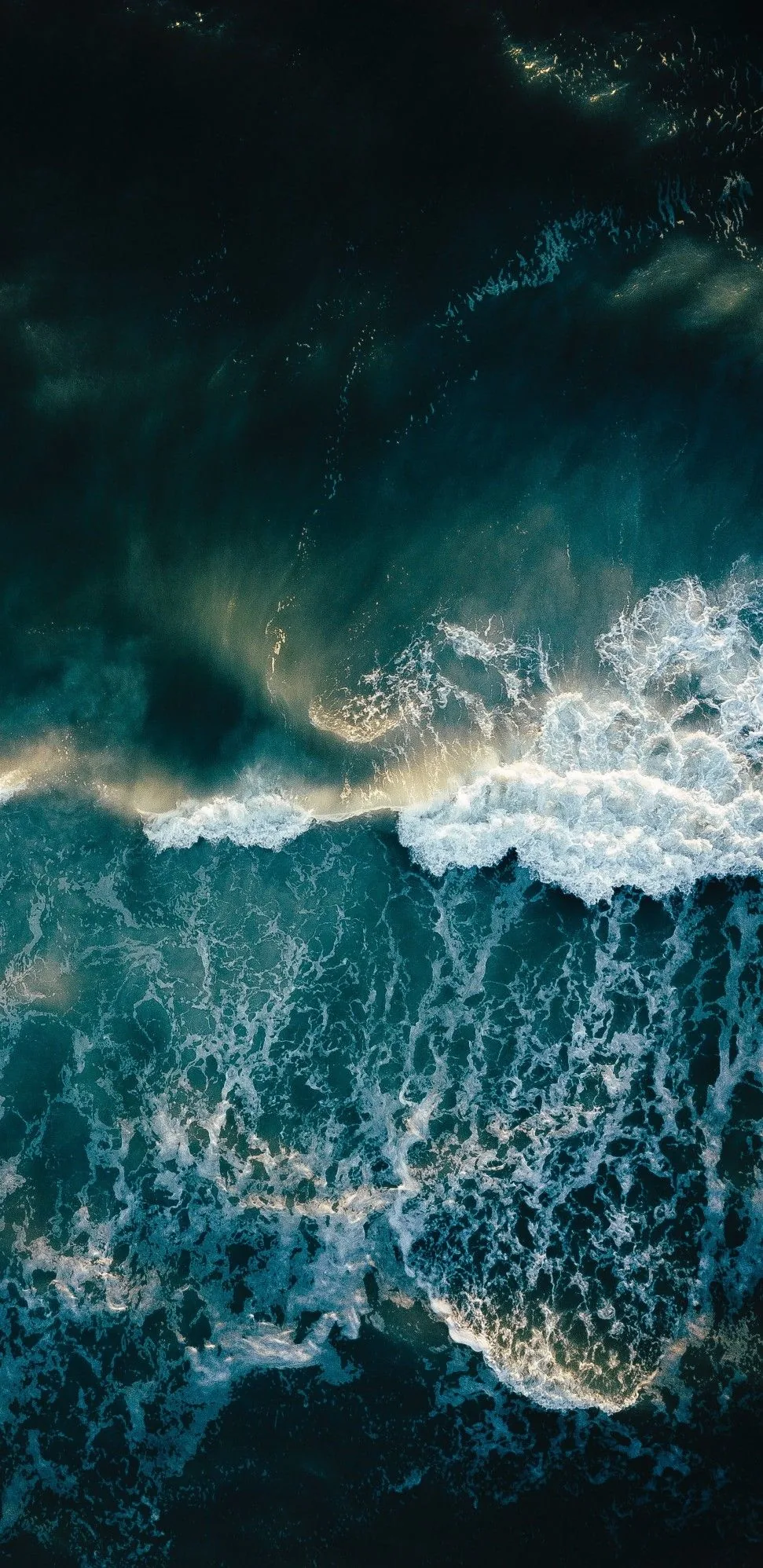 A dark aerial of storm swell, one broad band of white water tearing across the middle while the top corner fades to near black.