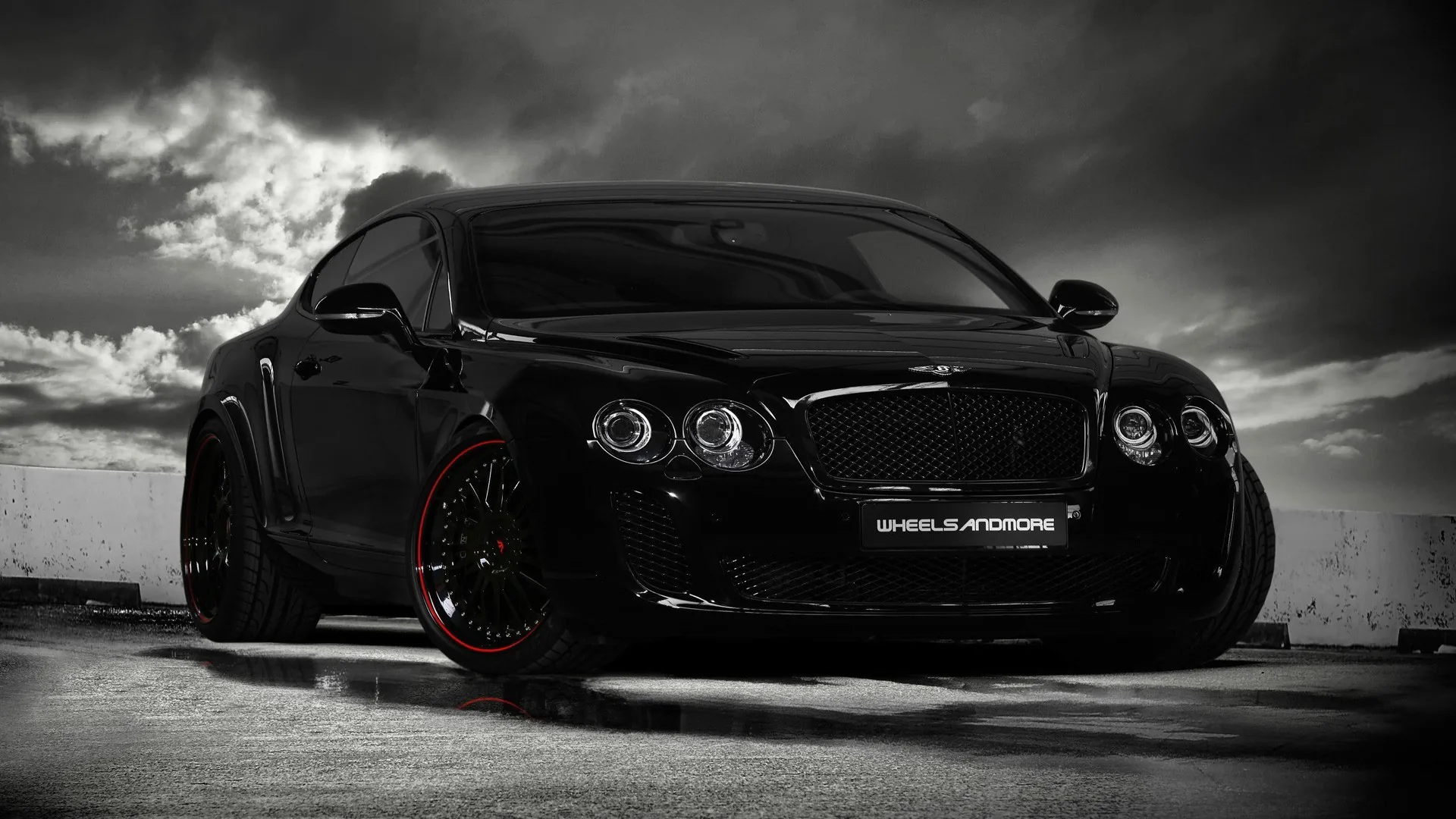 A stanced Continental GT shot low against dark cloud, its blacked-out mesh grille and quad headlamps sharp while red split rims sit under the arches.