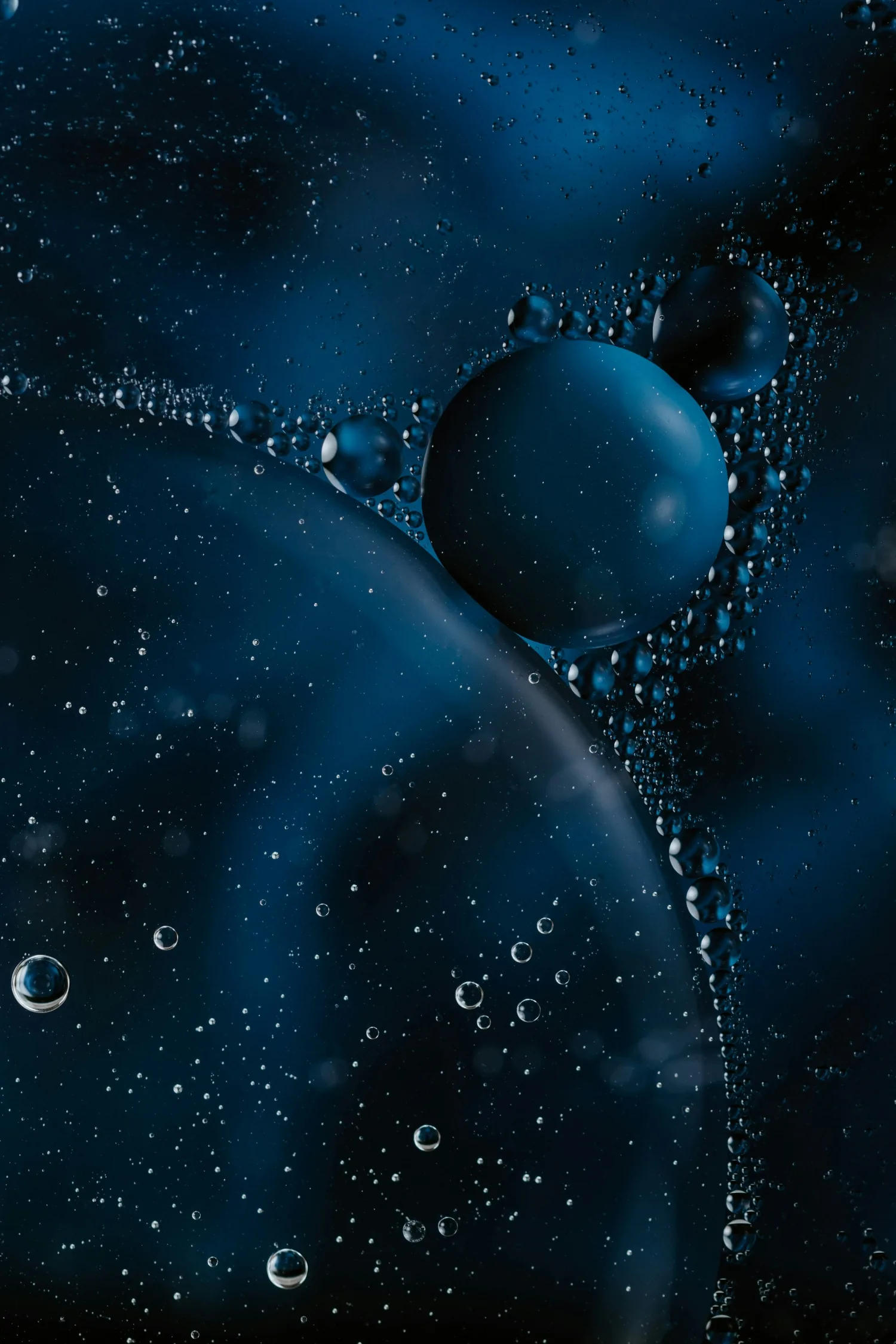 A macro shot of oil beading on water, one large teal sphere at the centre surrounded by smaller bubbles catching a cold rim light.