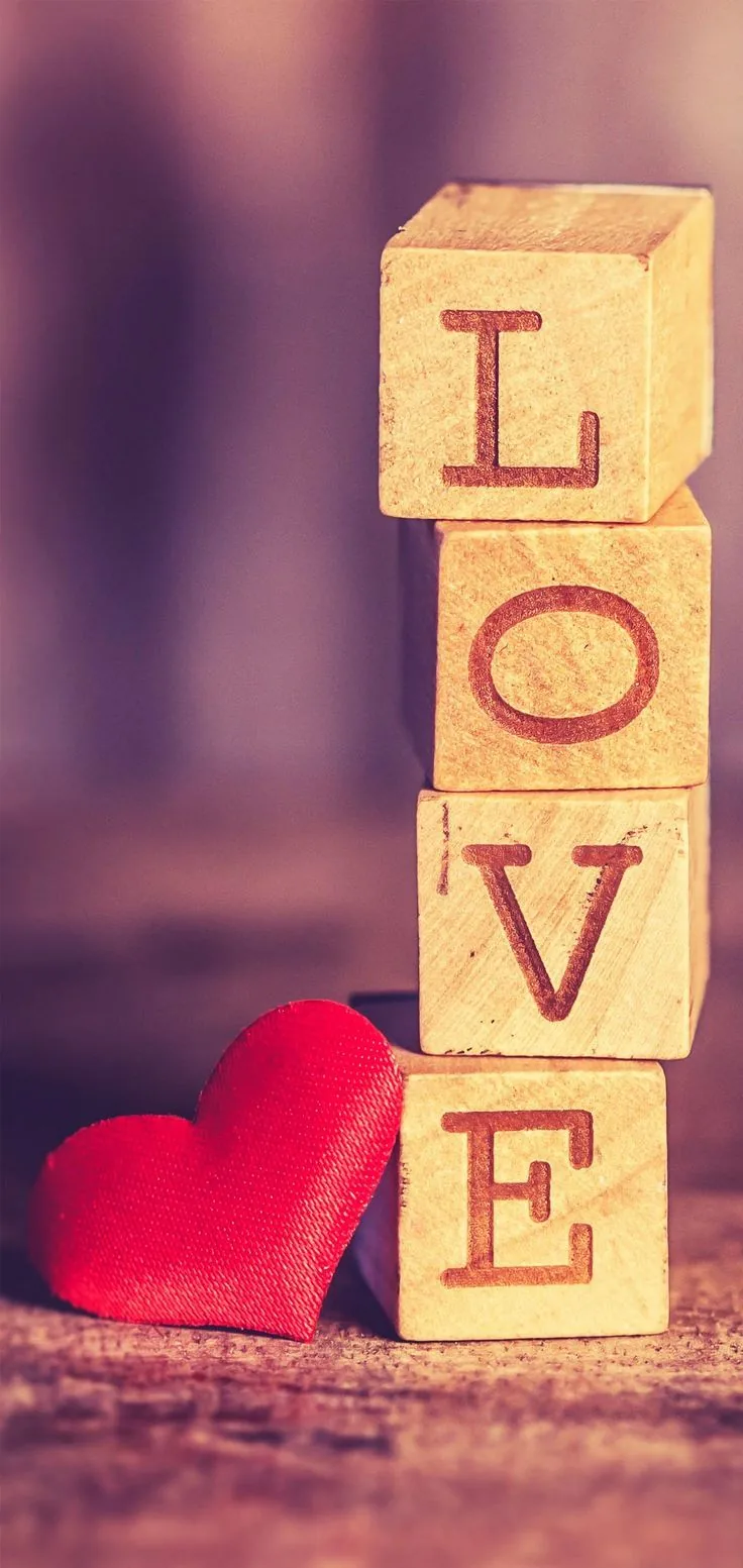 Four wooden alphabet blocks stacked into the word Love, a plush red heart leaning against the base on warm blurred wood.