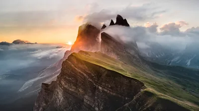Grass clings to a near-vertical ridgeline that drops into sunset fog, the low sun flaring in a notch between the rock teeth.