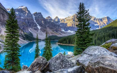 Turquoise glacial water stretches to a wall of grey summits, sunlit boulders and spruce in the foreground giving the shot its depth.