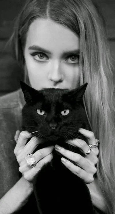 A long-haired girl holds a black cat up to her chin, both pairs of eyes on the camera; her ringed fingers frame the cat's face.
