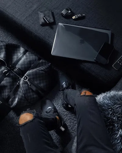 Seen from above, ripped jeans, leather gloves, a checkered duffel and a laptop spread across dark bedding, small keys and a wallet along the top.