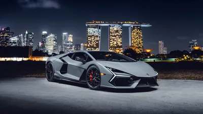 The hybrid V12 sits low on a waterfront lot at night, hotel towers and office lights stacked across the water behind its rear wing.