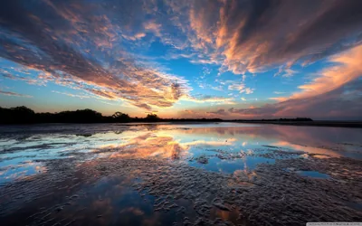 Sunset clouds and blue sky repeat in the wet sand of a tidal flat, the horizon splitting the frame into two mirrored halves.