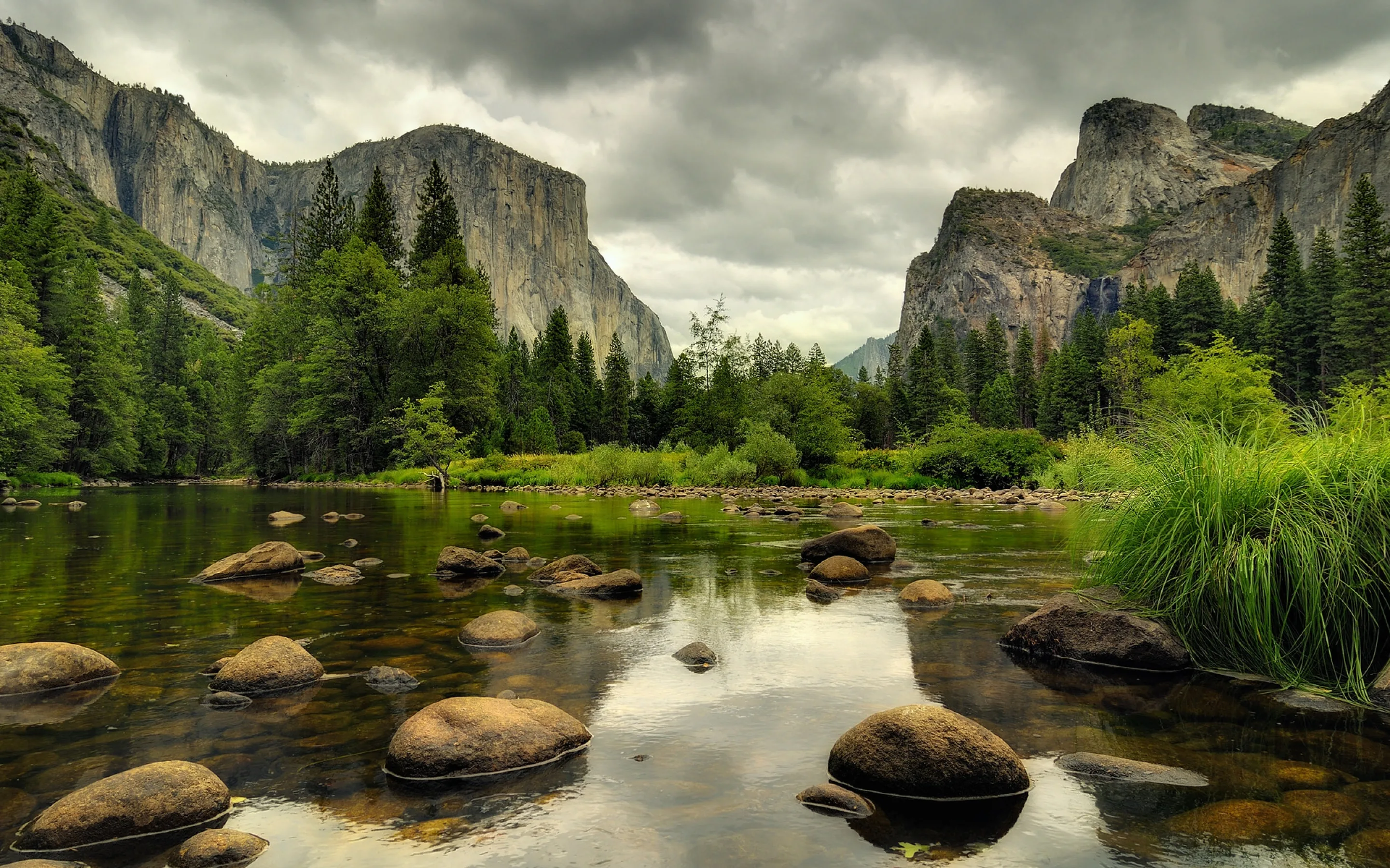 Granite walls rise on both sides of the Merced River, boulders leading in from the bottom edge; overcast light keeps the scene soft with no harsh shadows.