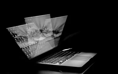 Translucent panels lift off a laptop screen in a black room, the keyboard catching the only strong light.