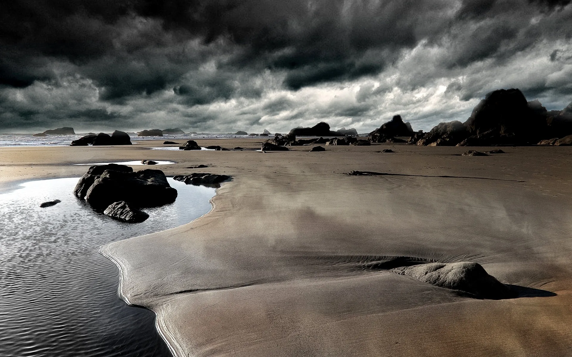 Heavy grey cloud presses down on a wet beach at low tide, scattered rocks and shallow pools catching what light gets through.