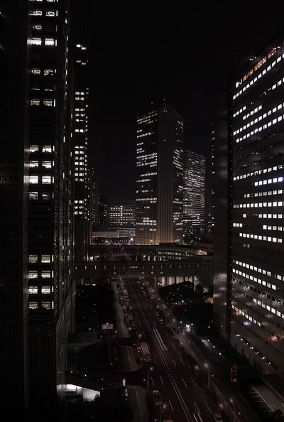 Shot from a high window: glass towers with scattered lit floors on both sides, and far below a multi-lane road smeared with red and white traffic trails.