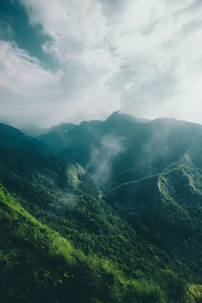Steep jungle-covered ridges climb into drifting mist under a bright white sky, the hazy upper third leaving clear space for a phone clock.