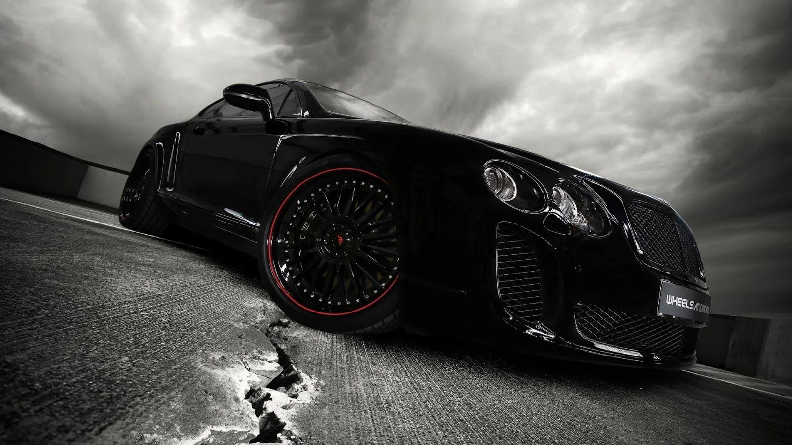 Low angle on a black Bentley with deep red-lipped wheels, heavy grey cloud stacked above the road and a small pool of water catching light near the front tyre.